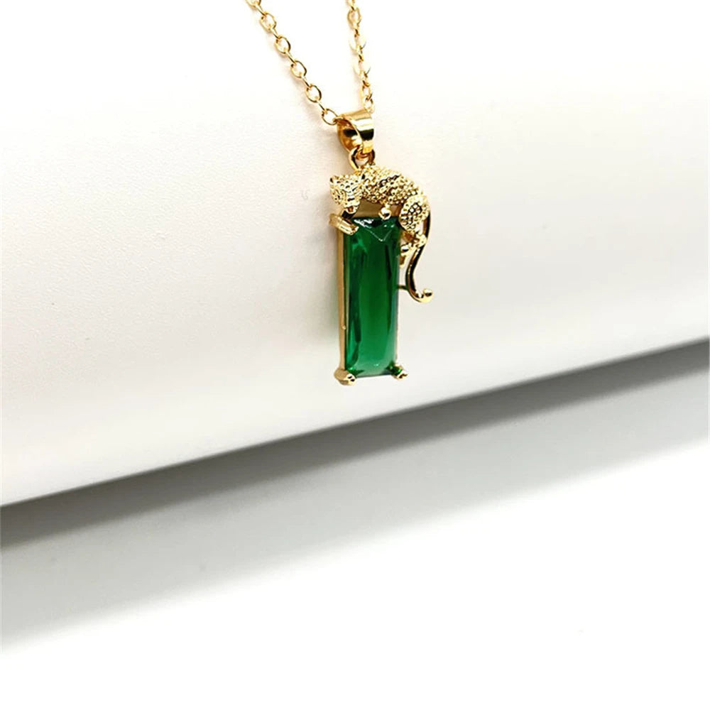 Colar dourado com pingente de leopardo segurando cristal verde retangular em fundo branco minimalista.