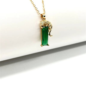 Colar dourado com pingente de leopardo segurando cristal verde retangular em fundo branco minimalista.