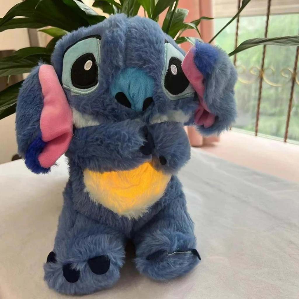 Pelúcia do Stitch azul com barriga amarela iluminada em superfície clara com plantas ao fundo.