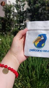 Mão segurando uma bolsa de tecido personalizada com o logo da Pelicano Azul, ao lado de uma pulseira vermelha com medalha dourada. Pelicano Azul.