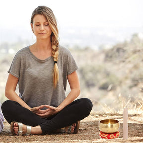 Mulher meditando ao ar livre em posição de lótus com uma tigela tibetana dourada ao lado, em um ambiente natural e tranquilo. A tigela está colocada sobre um suporte vermelho ornamentado, simbolizando a paz e o equilíbrio durante a prática meditativa.