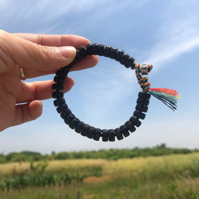 Pulseira Budista de contas de concha esculpida, com detalhes tibetanos e cordão multicolorido, sendo segurada contra o céu em um campo aberto, Pelicano Azul.