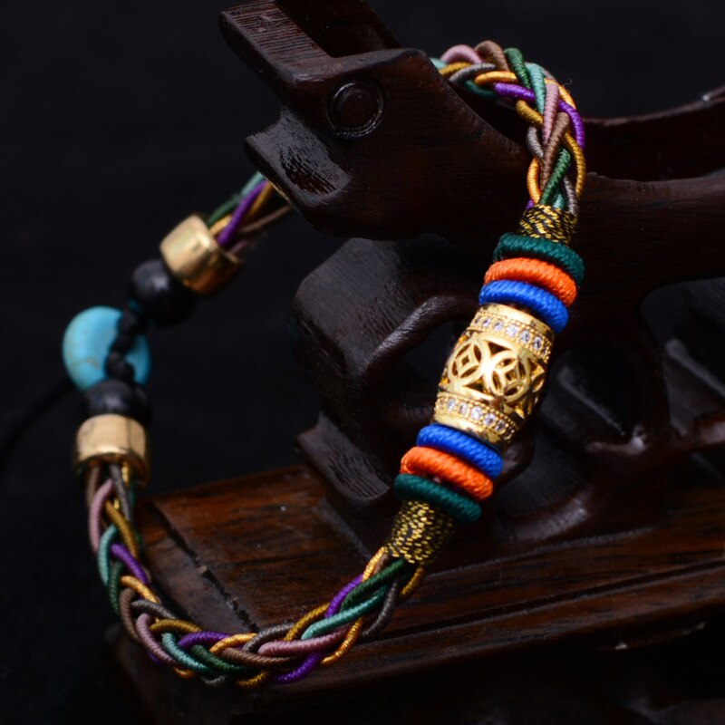 Pulseira Tibetana com 8 fios trançados em cores vibrantes, detalhe central dourado e pedras de turquesa, simbolizando saúde e prosperidade, Pelicano Azul.