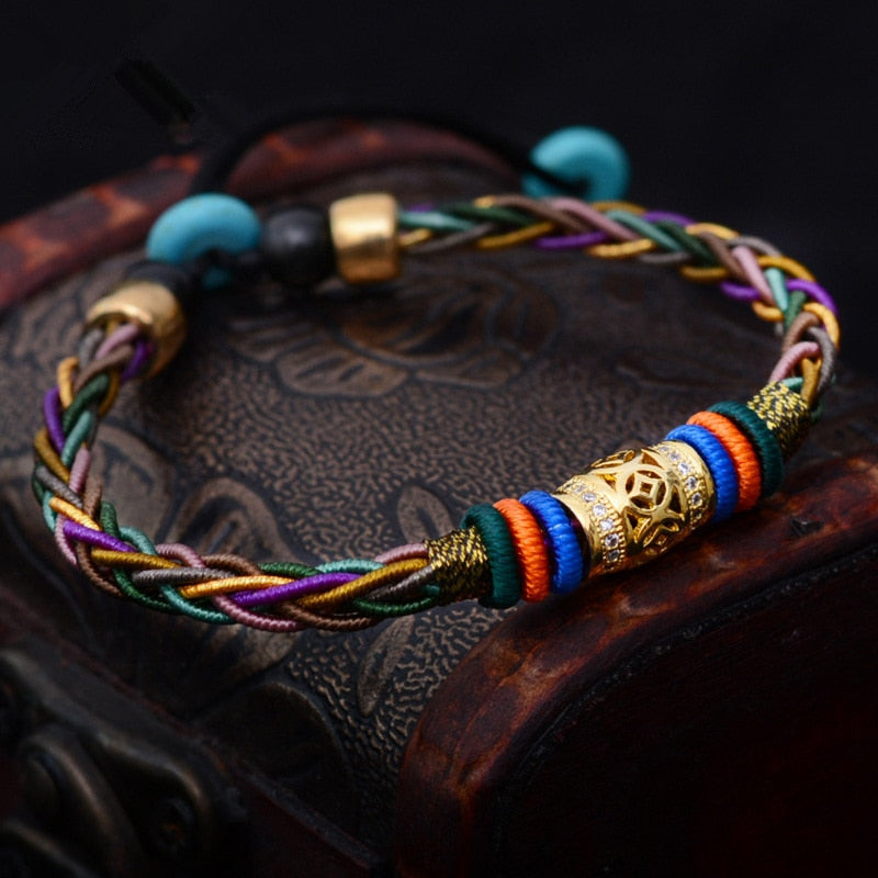 Pulseira Tibetana de 8 fios trançados com detalhe dourado central e pedras turquesa, representando saúde e prosperidade, Pelicano Azul.