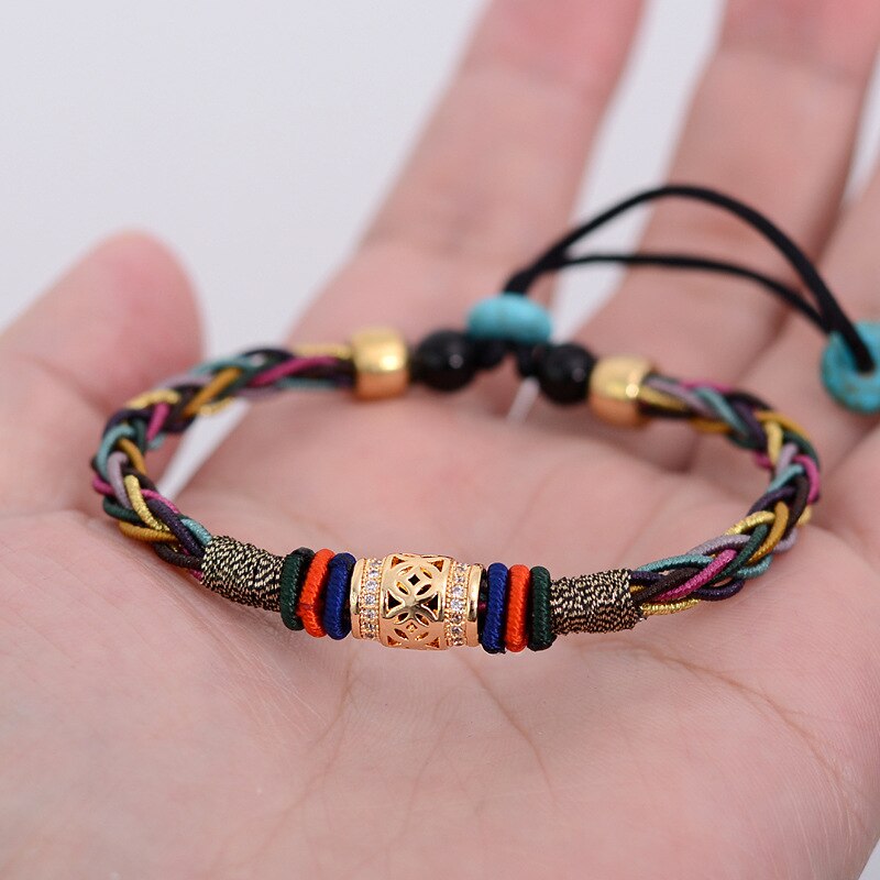 Pulseira Tibetana de 8 fios coloridos trançados com detalhes dourados e pedras de turquesa, símbolo de saúde e prosperidade, sendo mostrada na palma de uma mão.