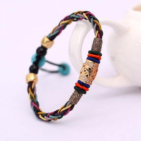 Pulseira Tibetana de 8 fios coloridos trançados com detalhes dourados e pedras de turquesa, símbolo de saúde e prosperidade, exibida em um suporte decorativo de fundo branco.