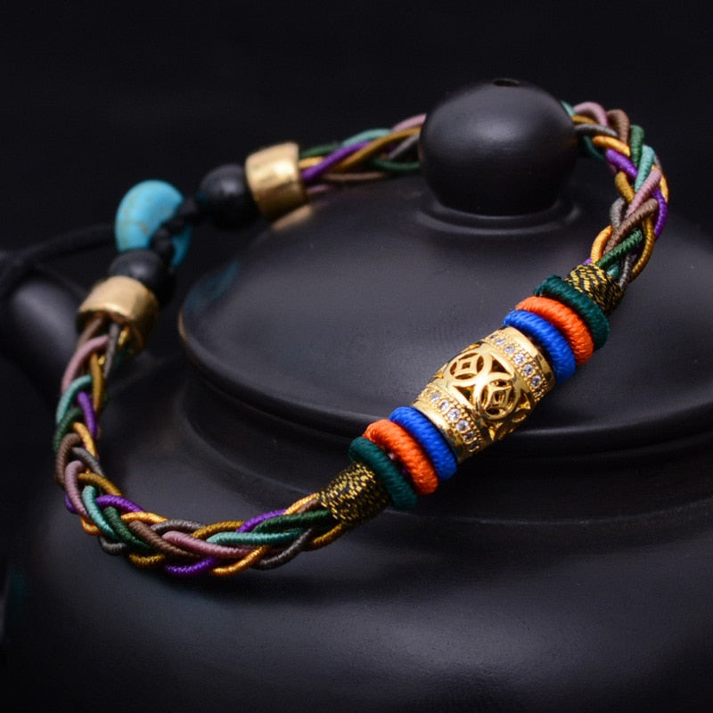 Pulseira Tibetana trançada com 8 fios multicoloridos, com detalhes em ouro e pedras turquesas, simbolizando saúde e prosperidade, Pelicano Azul.