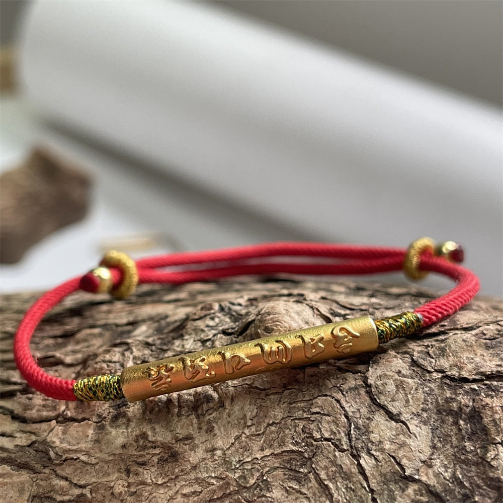 Pulseira Vermelha Tibetana Protetora com detalhe em dourado e inscrições tibetanas gravadas. Design elegante com cordão ajustável, ideal para proteção e equilíbrio espiritual. Pelicano Azul.